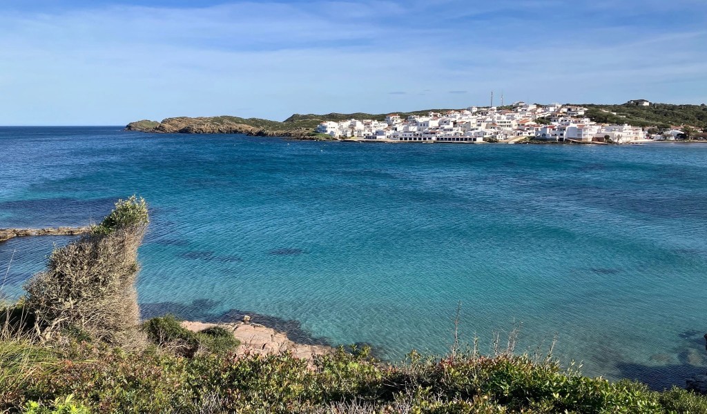 photo looking across blue-green water to a village of all-white, 1-2 story buildings on a peninsula on the opposite shore