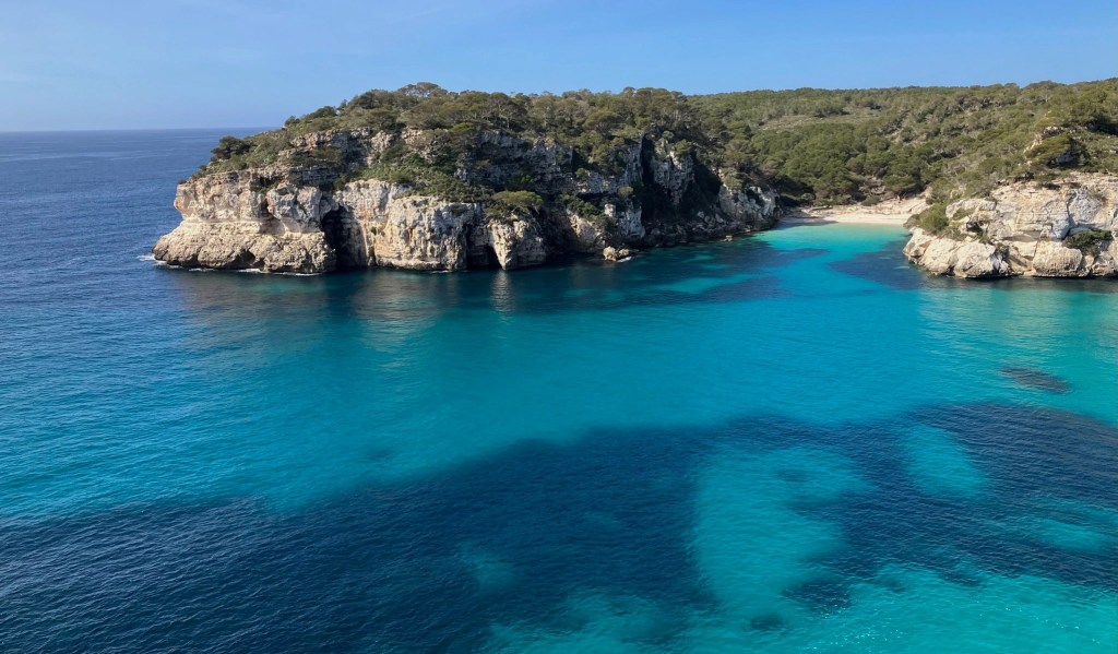 photo of turquoise and blue water in the foreground with rocky cliffs, green bluffs, and a small cove with a white-sand beach