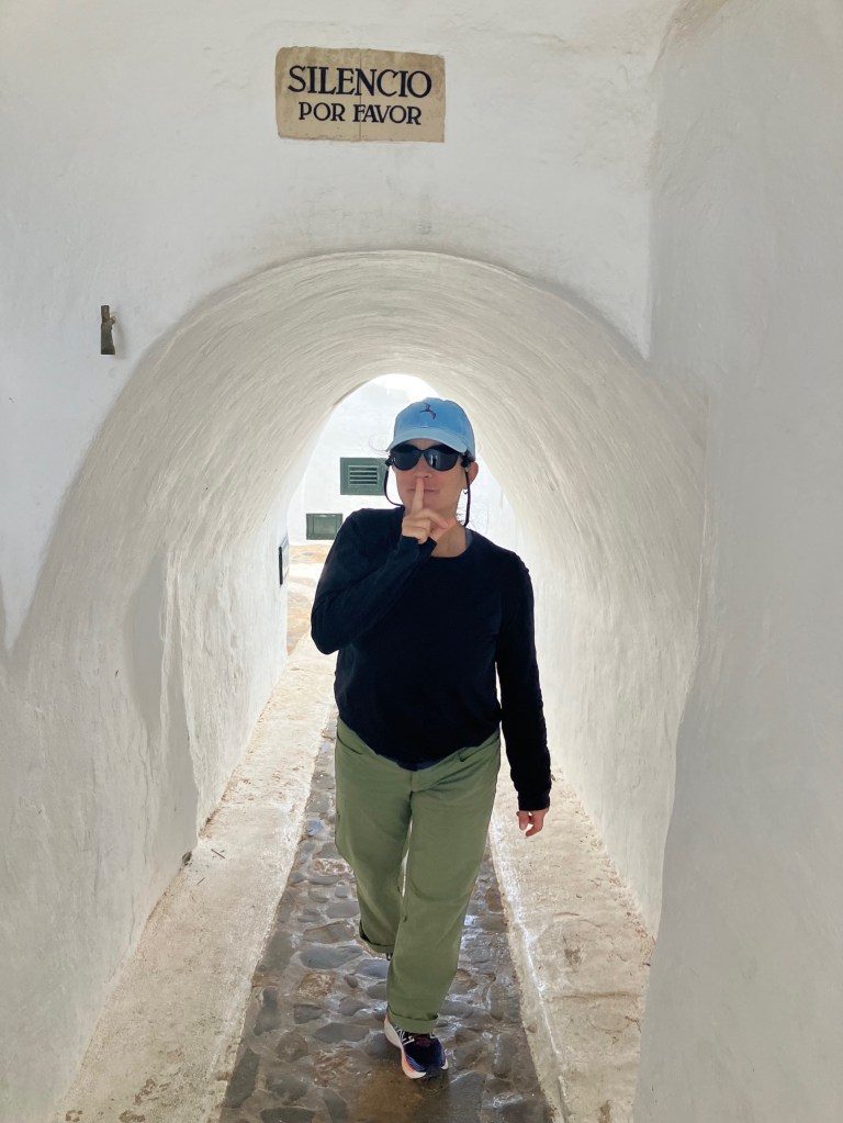 photo of a sign reading "Silencio Por Favor" above a white tunnel just big enough for the woman walking through it with her finger over her lips