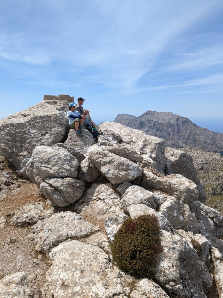 Photo of Al and Rachel perched on large, jumbled boulders overlooking a far-off valley with wispy clouds against a bright blue sky.