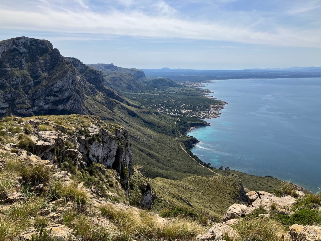 Photo of hills sloping toward aquamarine sea water with exposed rocks and vibrant green vegetation. A seaside town is off in the distance.