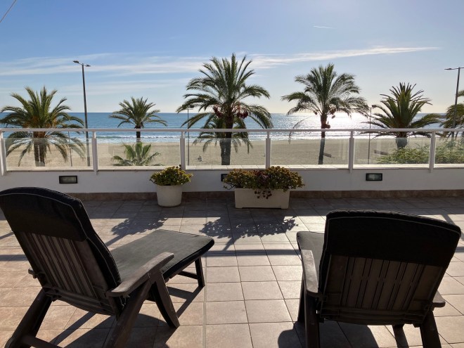 photo of two lounge chairs on a tile deck facing a row of palm trees with sandy beach and blue ocean beyond, and the sun casting a stream of white light on the water