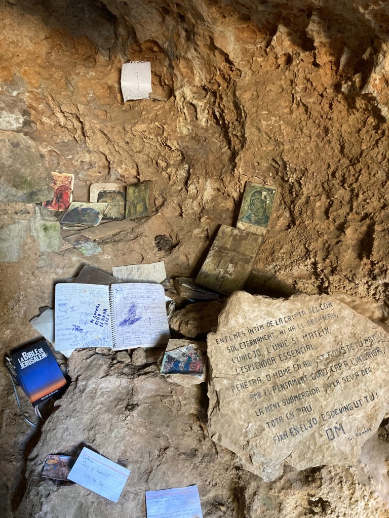 Photo of an informal shrine inside a stone cave carved from the rock with crude tools. The shrine has small pictures, a notebook, bible and handwritten cards scattered about on the rock.