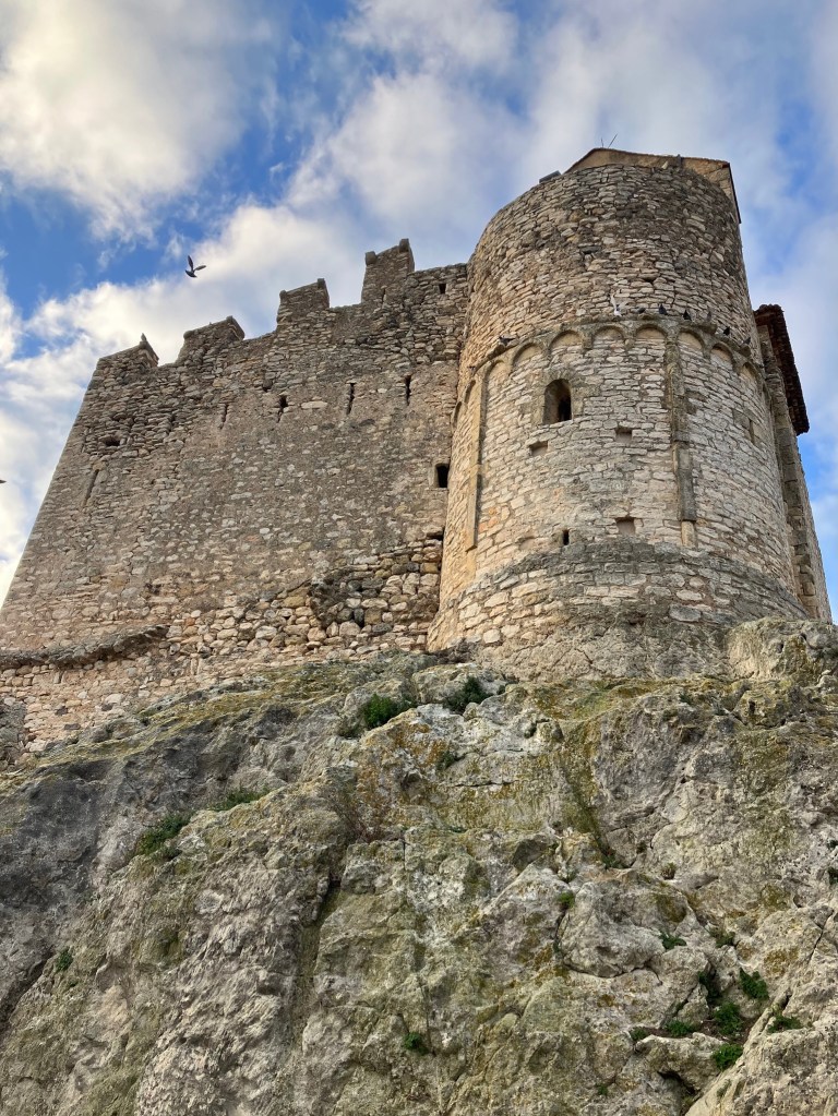 photo of a stone castle wall and tower on top of a rock cliff with blue sky and clouds above