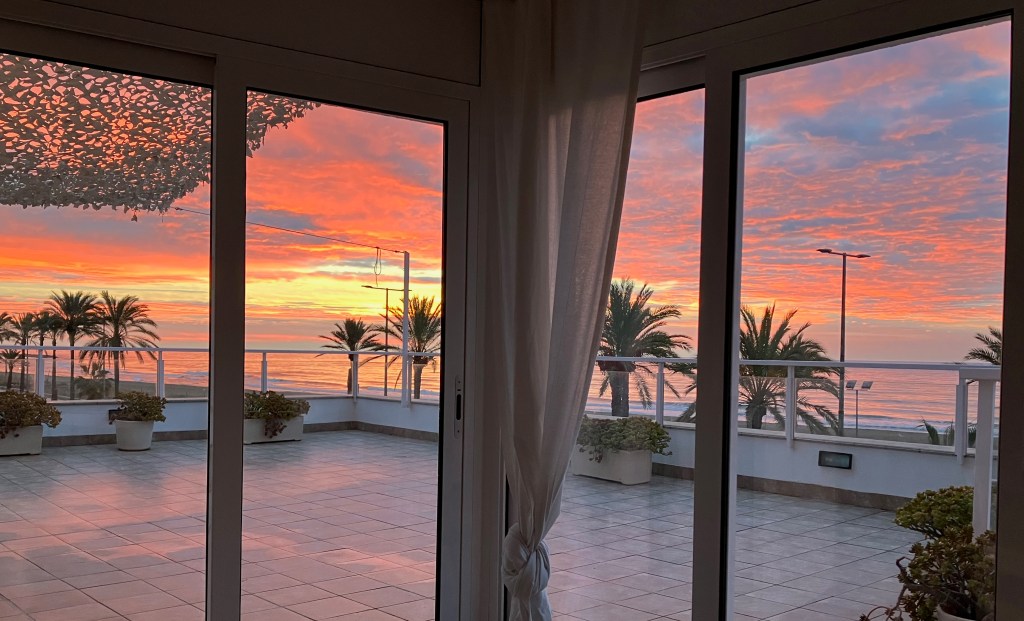 photo of sliding glass doors meeting in a corner overlooking a tile deck, with palm trees and a red-orange sunrise beyond