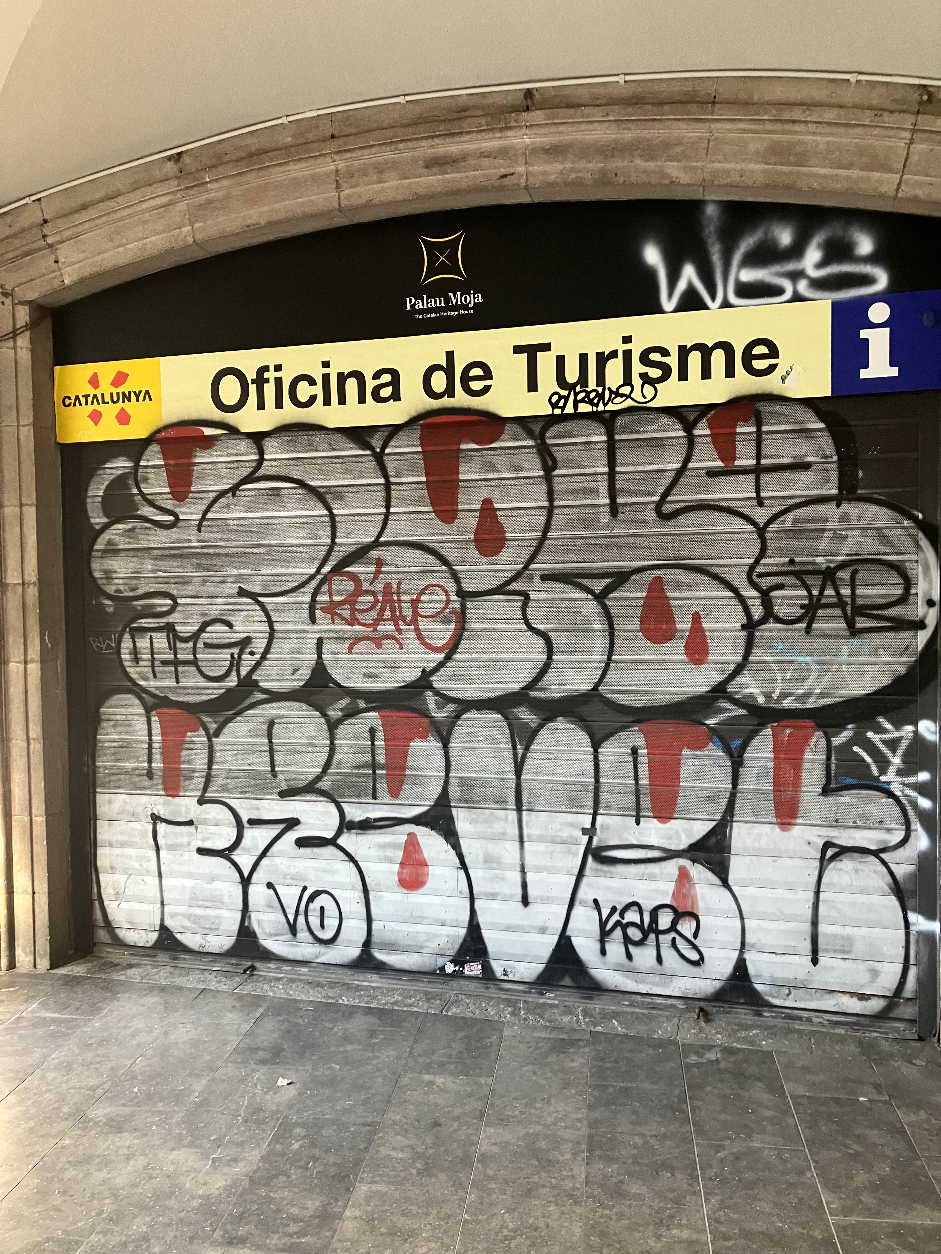 photo of a garage door with a sign "Catalunya Oficina de Turisme" above it and silver, black, and red graffiti letters spray-painted on it