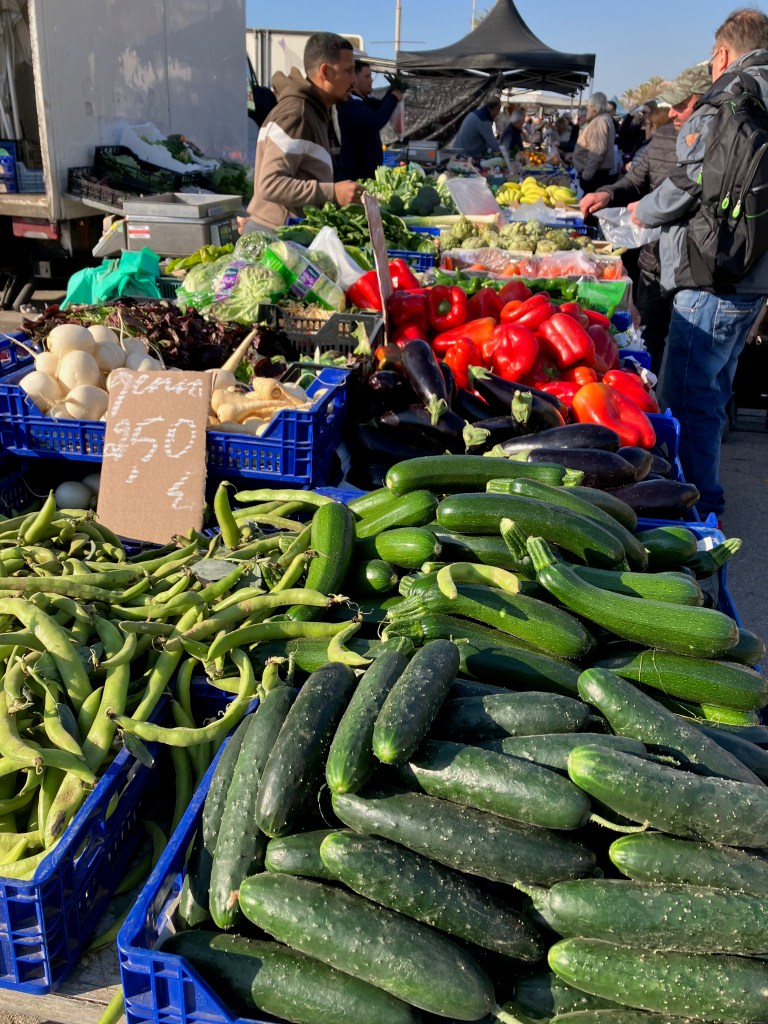 photo of an outdoor vegetable stand with cucumbers, beans, zuchini, and red peppers in the foreground and buyers talking with a seller in the background