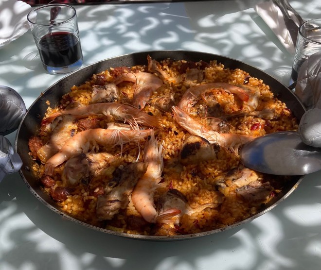 photo of a circular pan filled with brown-orange rice, chunks of browned meat, and topped with six large pink shrimp, sitting on a white table with a glass of wine and a serving spoon.