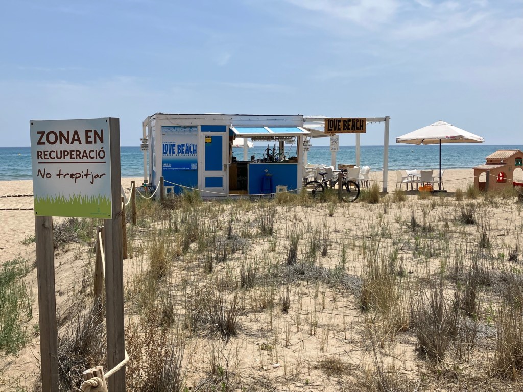 photo of a beach with a sign in the foreground saying "Zona en Recuperació" next to a roped-off area with sand and beach grass. Behind it is a cabana bar called "Love Beach" and an umbrella with chairs beneath it