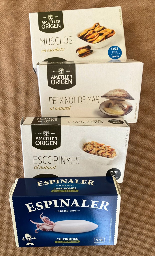photo of four boxes of tinned fish on a brown tabletop. Labels on the boxes are in Catalan: musclos (mussels), petxinot de mar (clams), escopinyes (snails), and chiprones (squid).
