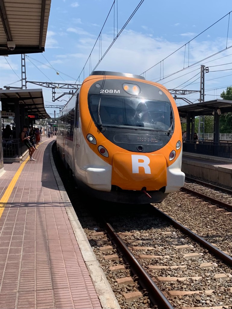 photo of the front of a train painted orange with a large round windshield and a capital R on parallel train tracks by a brick platform.