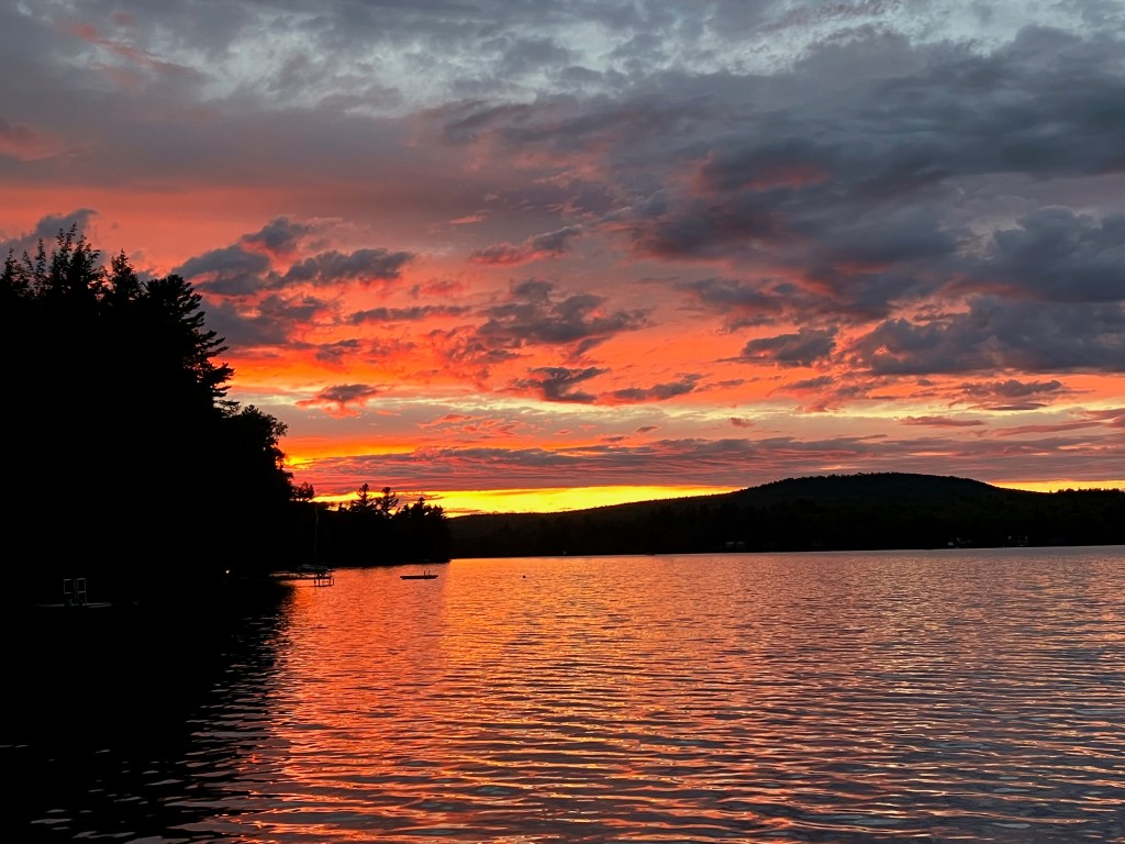 photo of deep red and orange clouds with bright yellow glow along a horizon lined by shadowy hills and trees, with a rippled lake in the foreground