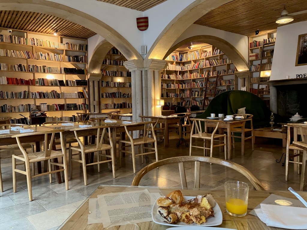 photo of a dining area with pastries and orange juice on a table in the foreground and shelves full of books in the background.