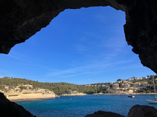 photo looking out of the mouth of a cave onto a blue harbor with two boats, yellow sandstone cliffs, a small hill with green trees, and a village with white and brown buildings rising up from the waterside.