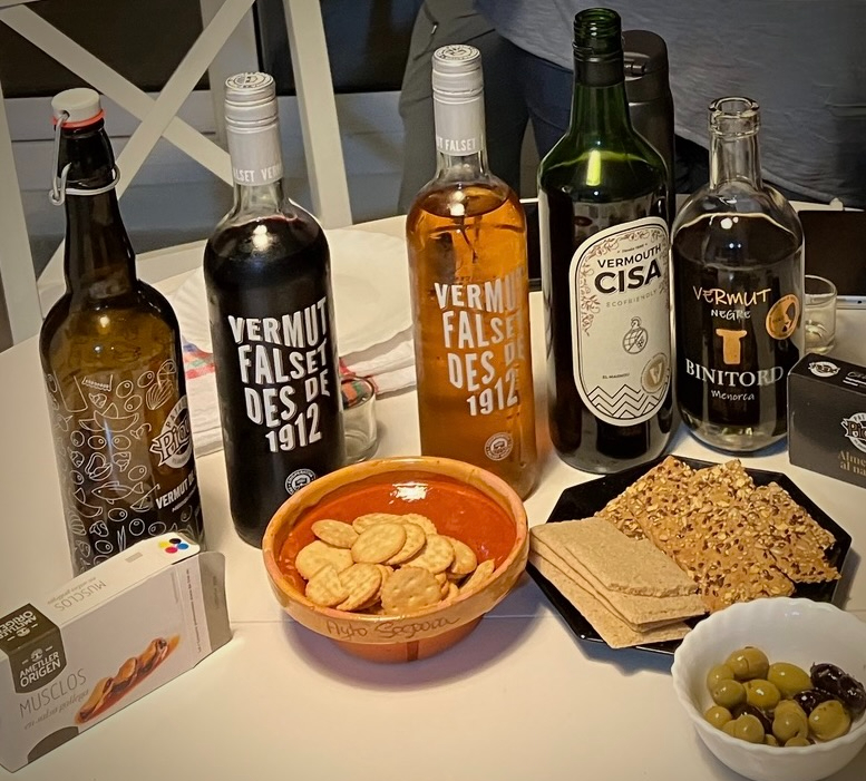 photo of five different bottles of vermouth lined up on a table with tinned mussels, crackers in a bowl and a plate, and a bowl of olives