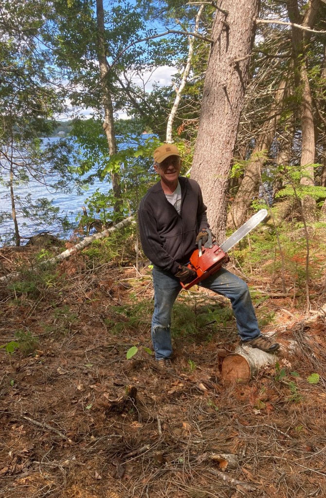 photo of a man holding a chain saw with a fallen birch tree at his feet, standing trees behind him, and brown scrub brush on the ground, with a lake in the background
