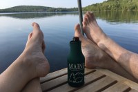 photo of two sets of feet crossed at the ankles, resting on a wooden table with a beer in between, wrapped in a green coozie labeled "Maine", with a glassy lake in the background reflecting green tree-covered hills.