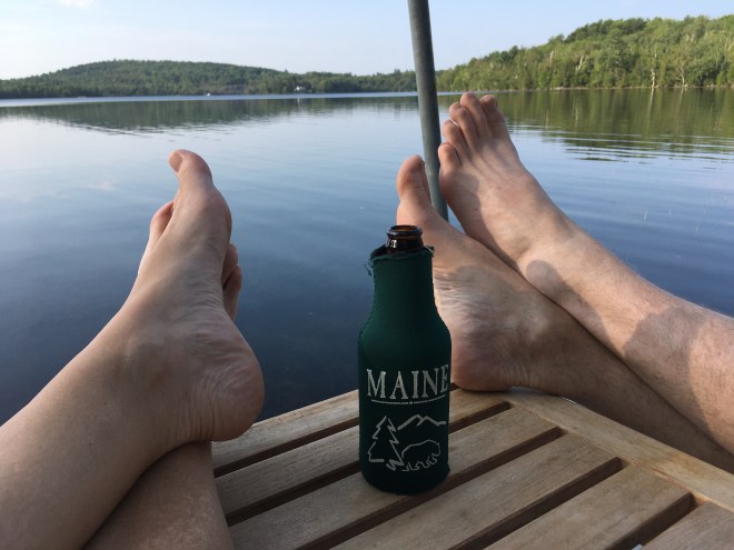photo of two sets of feet crossed at the ankles, resting on a wooden table with a beer in between, wrapped in a green coozie labeled "Maine", with a glassy lake in the background reflecting green tree-covered hills.