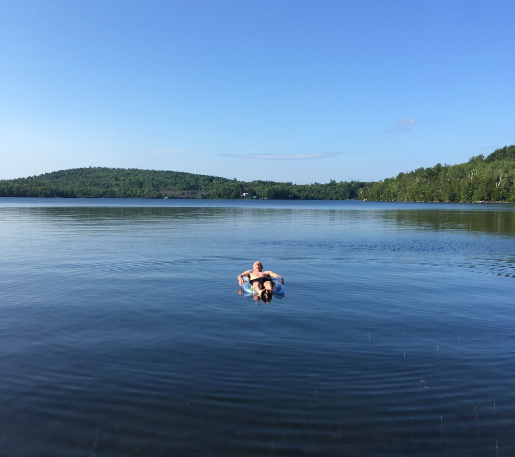 photo of a man reclined on a float in a quiet lake with blue sky above and rolling green hills behind