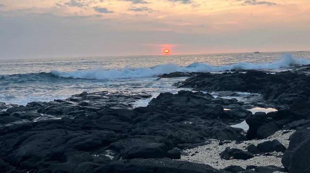 photo of a rocky shoreline with waves breaking on it and the sun setting in the background.