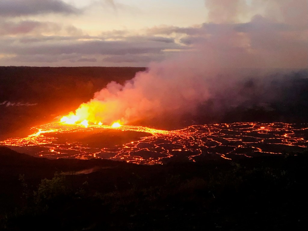 photo of bright yellow-orange flames bursting from a dark, wide crater and orange rivulets of lava swirling across the flat, dark landscape
