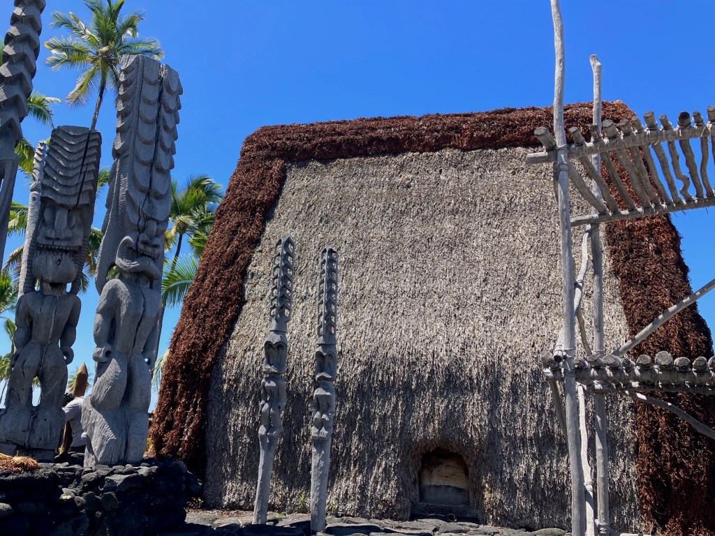 Photo of a gray and brown thatched building with carved wooden statues depicting Hawaiian gods.