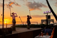 photo of two men wearing breach cloths on a stage, blowing conch shells and holding torches, with a sunset on the ocean behind them