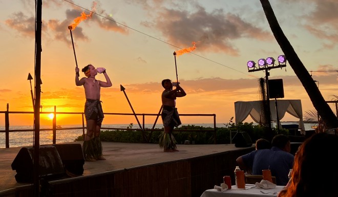 photo of two men wearing breach cloths on a stage, blowing conch shells and holding torches, with a sunset on the ocean behind them