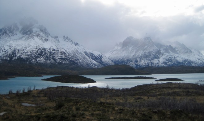 photo of craggy, snow-draped, dark mountains with their tips in a bank of clouds and a lake with small rounded islands in the foreground