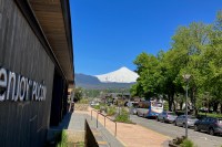 photo of a street and sidewalk with a white-covered, cone-shaped volcano in the background against a blue sky, and the side of a building along the left with a sign saying "enjoy Pucón" on it