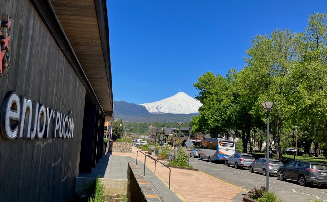 photo of a street and sidewalk with a white-covered, cone-shaped volcano in the background against a blue sky, and the side of a building along the left with a sign saying "enjoy Pucón" on it