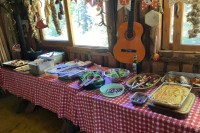 photo of a long table with red and white checked tablecloths. 15 serving dishes sit on the table, holding salds, potatoes, stuffing, turkey and vegetables. In the background strings of herbs and dried peppers hang in large windows and a guitar hangs from the wall.s
