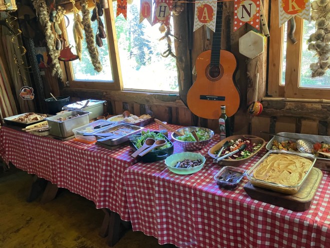 photo of a long table with red and white checked tablecloths. 15 serving dishes sit on the table, holding salds, potatoes, stuffing, turkey and vegetables. In the background strings of herbs and dried peppers hang in large windows and a guitar hangs from the wall.s