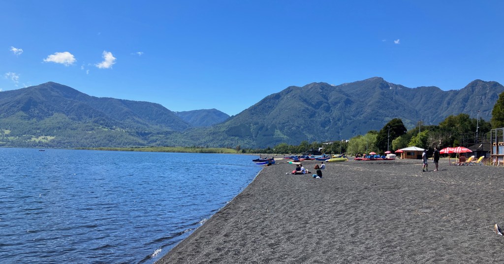 photo of a gray-black sandy beach on the rim of calm silvery-blue water and green, jagged mountains and blue sky in the background. Four or five people stand and sit on the beach, and a row of multi-colored kayaks and umbrellas sit on the beach in the distance