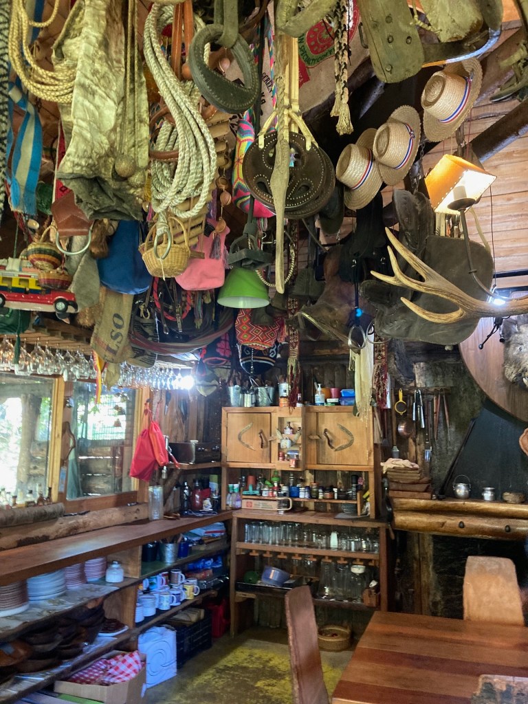 photo of a room whose ceiling is hung chock-a-block with hats, rope, fabric, baskets, burlap sacks, lamps, and antlers. Shelves and cabinets are equally jammed with jars, bottles, glasses, plates, cups, napkins, seasonings, and utensils.