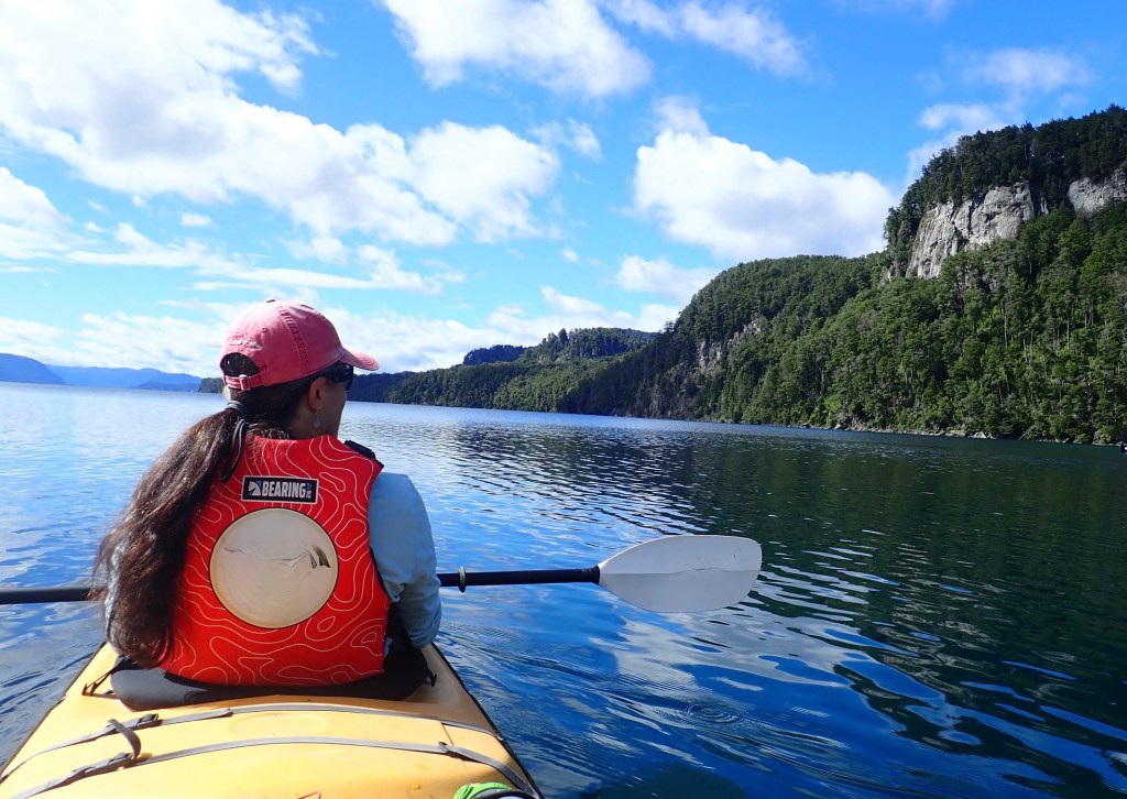 photo of a woman in the front of a yellow kayak, holding a paddle and wearing a red life vest and red ball cap with a long dark ponytail hanging down, looking across placid reflective waters at a gray cliff jutting out of a green forest