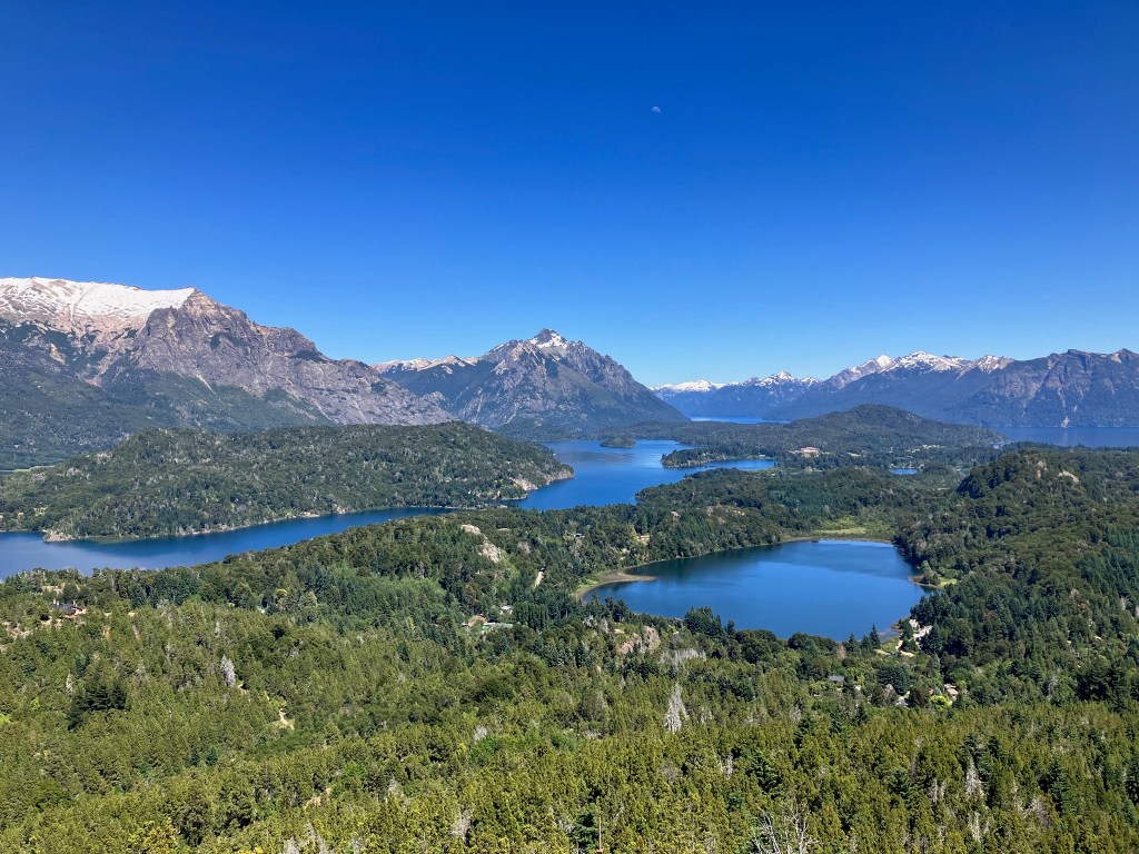 panoramic photo of a landscape with craggy snow capped mountains in the background and lakes of varied shapes and sizes surround by green forest in the foreground