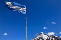photo of a blue and white flag of Argentina blowing in the wind at the top of a flagpole with a blue sky and snow-capped volcano in the background
