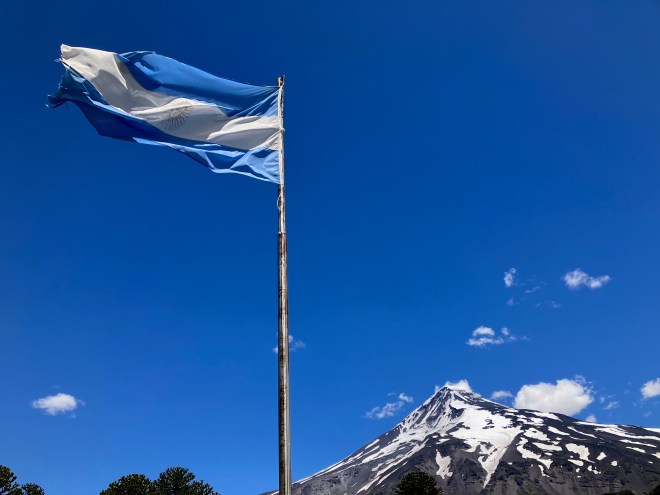 photo of a blue and white flag of Argentina blowing in the wind at the top of a flagpole with a blue sky and snow-capped volcano in the background