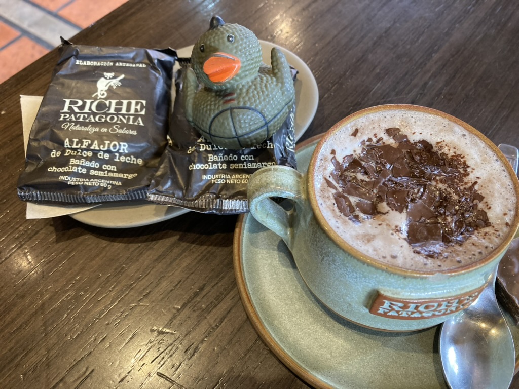 photo of (left to right) a RICHE Patagonia Alfajor in a brown wrapper, a dark green rubber duck with orange beak, and a green coffee mug with hot chocolate and melted chocolate pieces, all sitting on a dark wood table