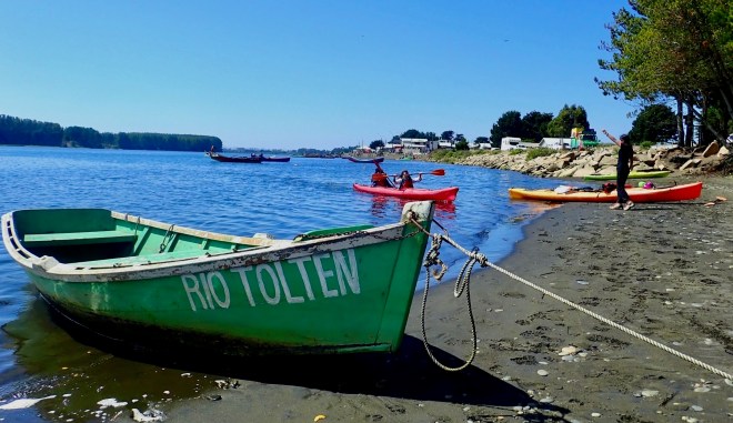 photo of a green wooden rowboat with RIO TOLTEN in white letters, resting on a dark sand beach. In the background is a wide blue river, a red kayak carrying two people lifting their paddles and an orange kayak on the beach with a person cheering next to it.