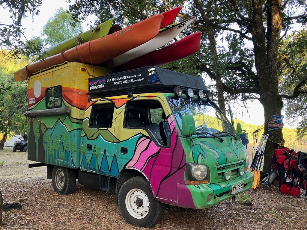 photo of a colorful truck, painted with a mural of green and purple mountains and red and orange sky, with 5 kayaks on top, parked between trees.
