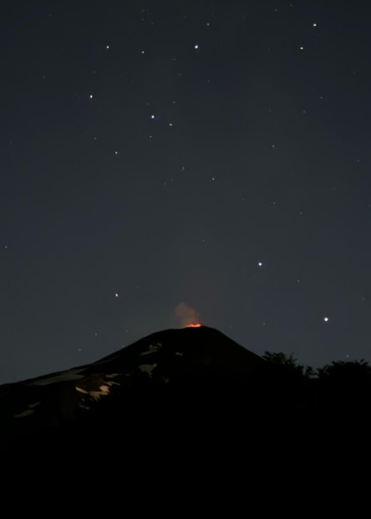 photo of the night sky, with white stars above, the silhouette of a cone-shaped volcano in the foreground and an orange glow emitting from the volcano's cone