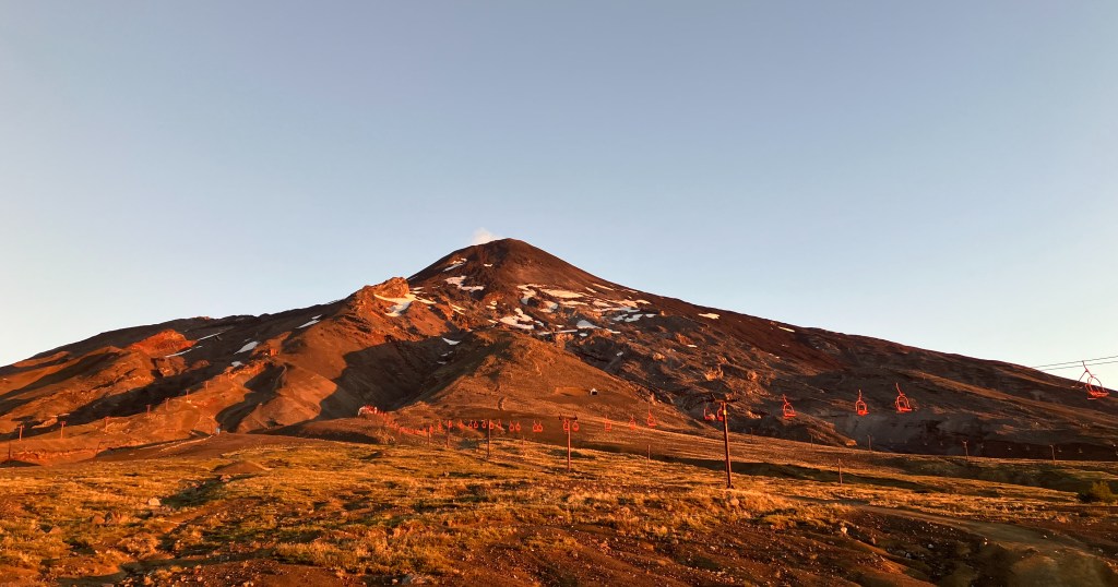 photo of a golden-brown, cone-shaped volcano with wisps of smoke emerging from th top, a few snow patches on the slopes, and a red chairlift along the bottom, with a dark blue sky overhead