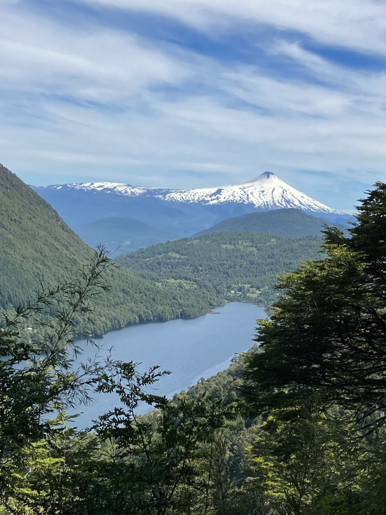 photo of a narrow blue lake set in a green valley with pine trees framing the foreground and a white, snow-covered, cone-shaped volcano in the background with wispy white clouds in a blue sky overhead