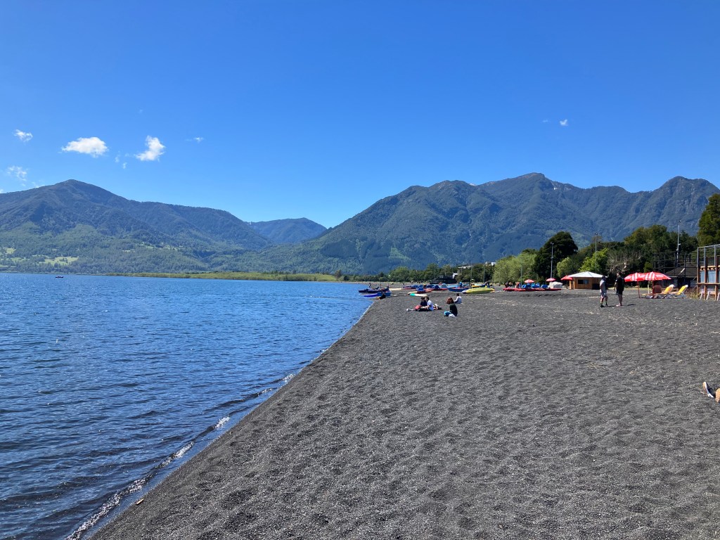 photo of a grey-black sand beach with still blue lake waters to the left, green hills to the rear, and a few people and tents scattered on the beach.