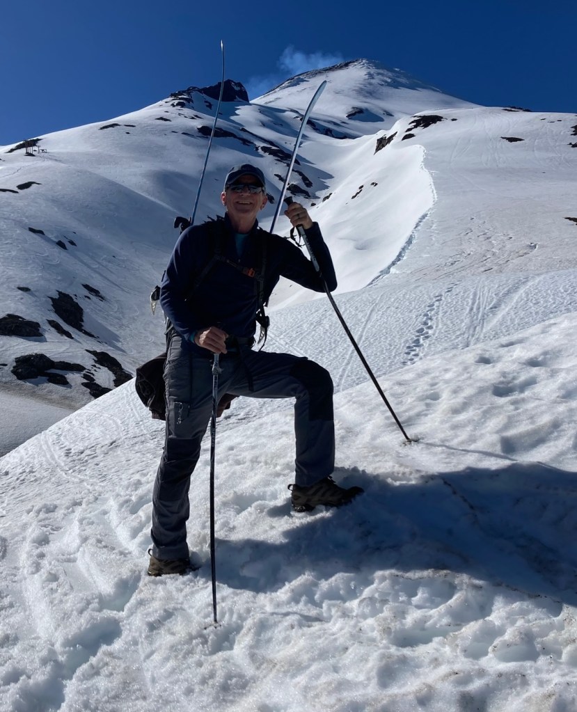 photo of a man stepping up a snow-covered slope with poles in his hands and skis on his back. Above and behind him are open, snowy expanses with a smoking peak at the top and blue sky behind.