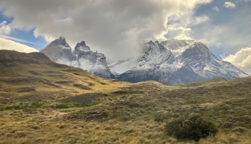 photo of a hill with golden grass and jagged gray mountains dusted with white snow in the background and clouds in the sky above