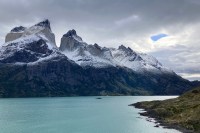 Photo of jagged brown mountain spires dusted with white snow above black cliffs and a glistening teal-green lake, with gray clouds and one small spot of blue sky above.