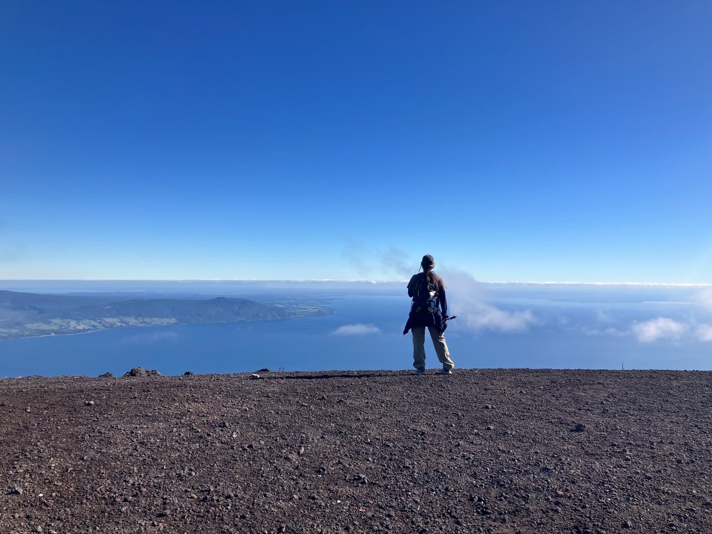 photo of a woman standing on flat gray gravel at the edge of a wide blue view of a lake and islands in the distance and blue sky overhead